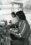 Native American man works on pottery while woman threads/sews in background